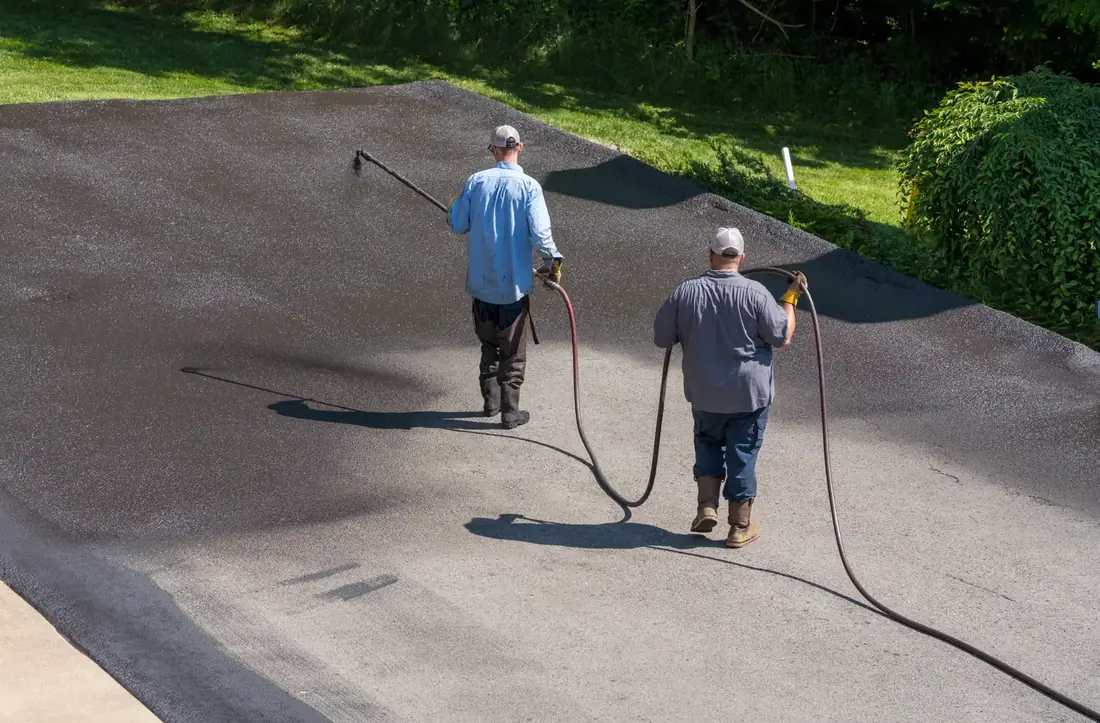 Two-person crew spray-applying asphalt sealcoat with a wand and hose, showing freshly sealed black pavement next to faded gray asphalt