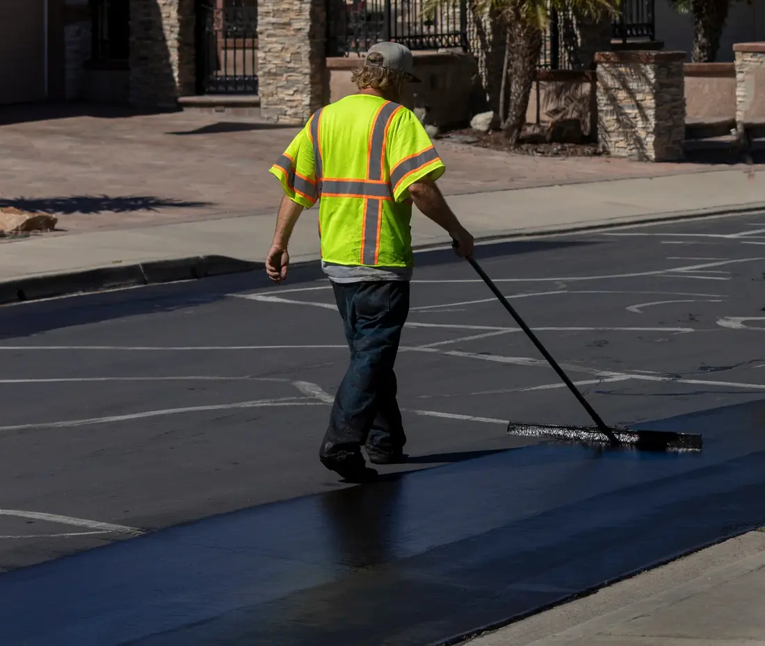 Worker in a safety vest applying asphalt sealcoat with a squeegee on a commercial surface