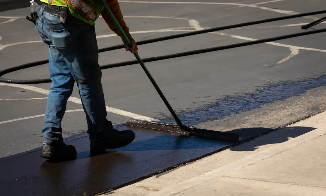 Worker in a safety vest spreading fresh asphalt sealcoat along a curb with a squeegee in a striped commercial parking lot